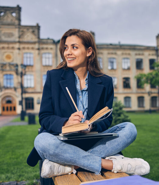Caucasian brunette student sitting with her laptop on knees while studying on the bench outside